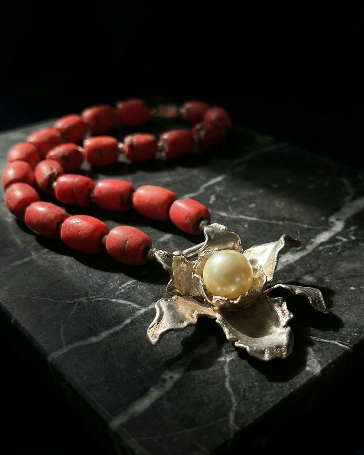 Coral bead necklace with a silver orchid clasp on black veined marble, lit chiaroscuro
